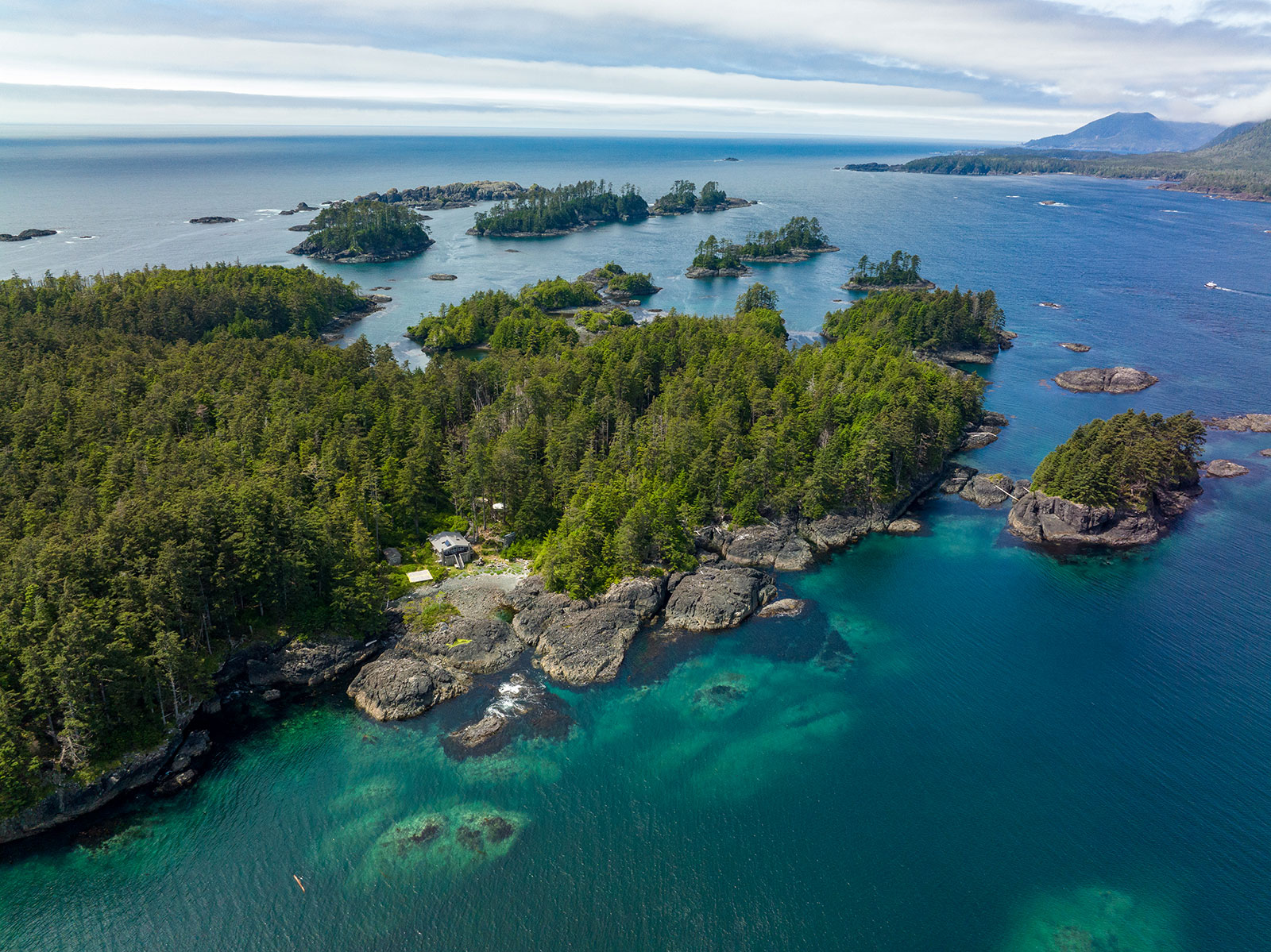 An aerial shot looks down at islands in Gwaii Haanas. The water is clear enough to see large underwater boulders and seaweed. Coniferous trees coat the land above the high-tide line.