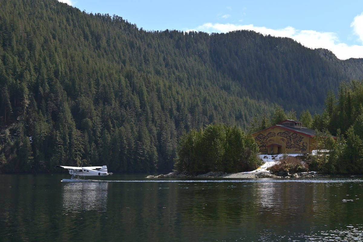 A floatplane lands on the water in front of the shoreline, which is home to a longhouse. 