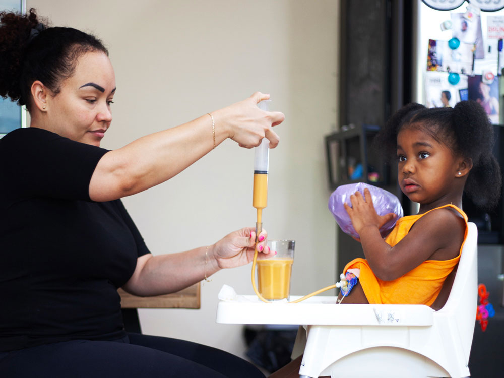 A woman fills a syringe with food for her daughter, who is seated in a high chair.
