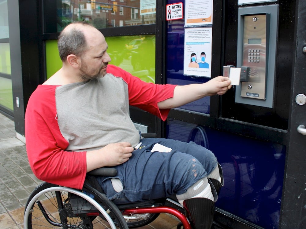 A man in a wheelchair uses a card to tap an electronic reader to gain access to a building.