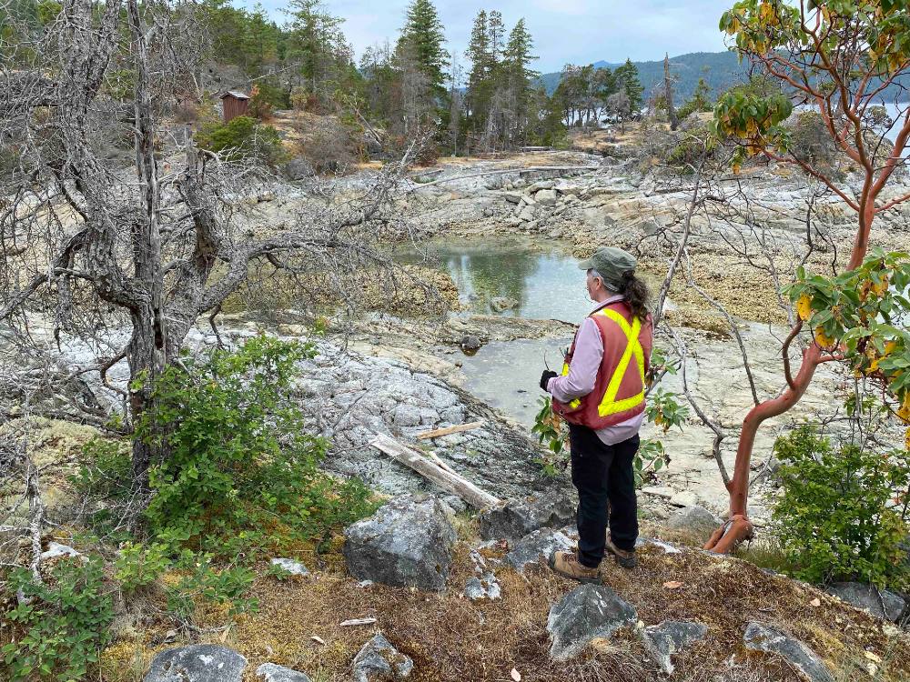 A woman wearing a hi-vis vest, hiking boots and gloves has taken her glasses off to better look at an area between two islands that is currently covered in Pacific oysters.