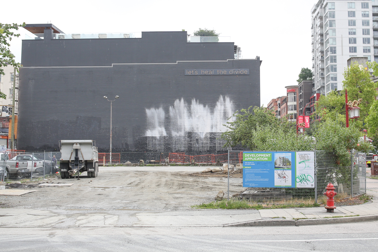 An empty piece of land with some dirt trucks. It is fenced off. On the fence is a bright blue design for a development application with a picture of a condo building on it. On the side of an adjacent building in the back are words in neon: “Let’s heal the divide.”