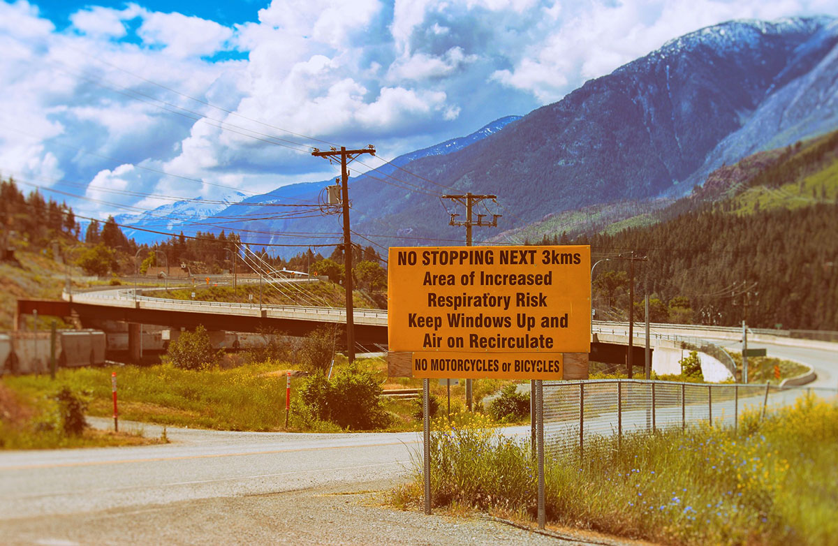An orange sign next to a road says “no stopping next three kilometres, area of increased respiratory risk, keep windows up and air on recirculate.” There is mountainous terrain in the background.