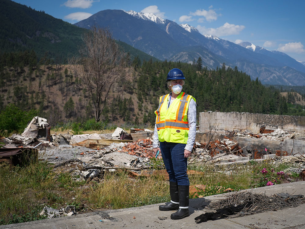 A woman wearing a yellow work vest, mask and hard hat stands in front of a demolished building. There are mountains in the background.