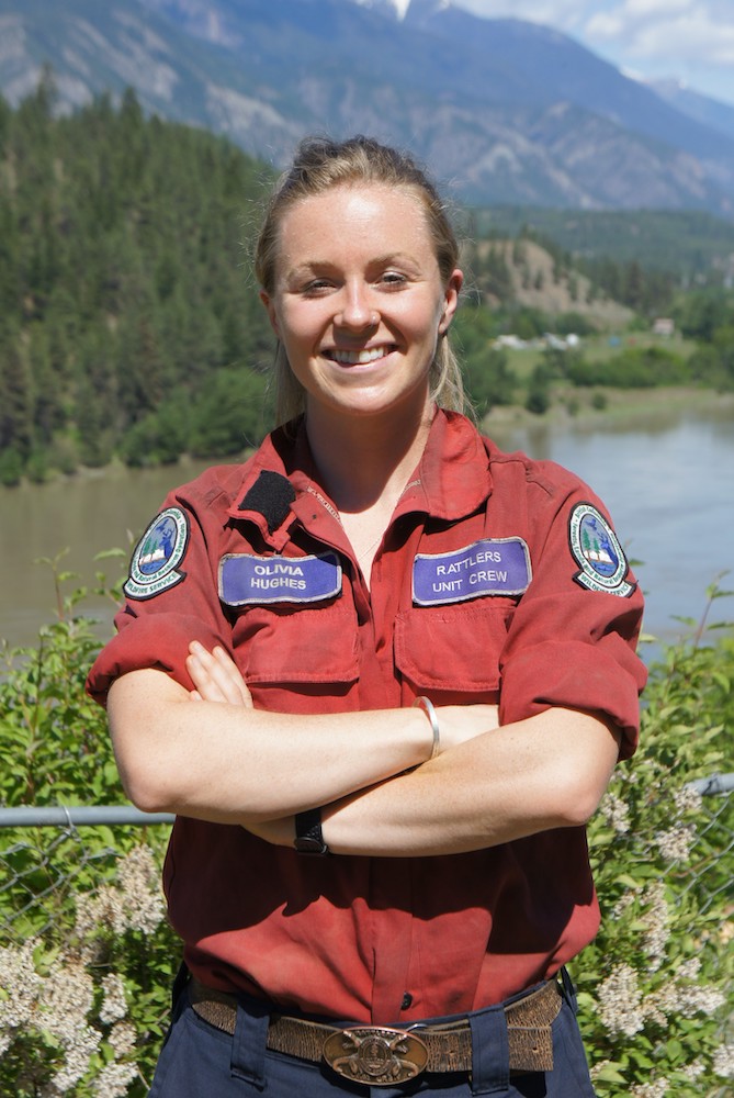 A young woman with blonde hair, wearing a red uniform, crosses her arms and smiles at the camera. Behind her is a river and mountainous terrain.
