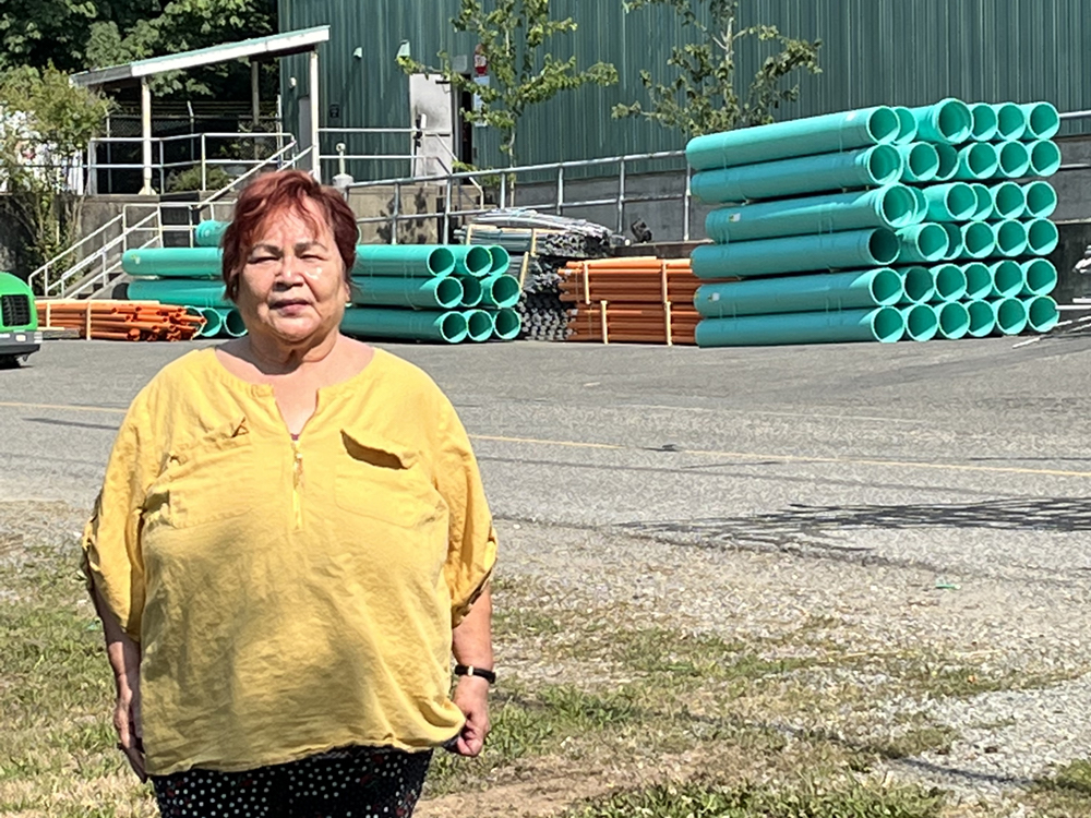 A woman stands on her driveway in front of a factory. There are PVC pipes on the ground.