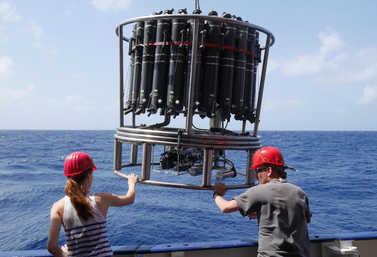 From behind, a woman and man maneuver a large, drum-shaped device hoisted on cables on the deck of a ship, with the sea behind.