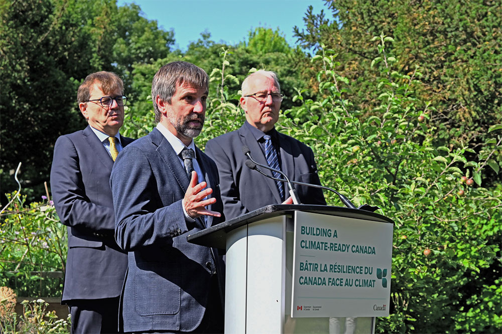 Federal Minister of Environment and Climate Change Steven Guilbeault stands at a podium, speaking, flanked by Minister of Emergency Preparedness Bill Blair to the right, and Jonathan Wilkinson, Minister of Natural Resources, to the left.