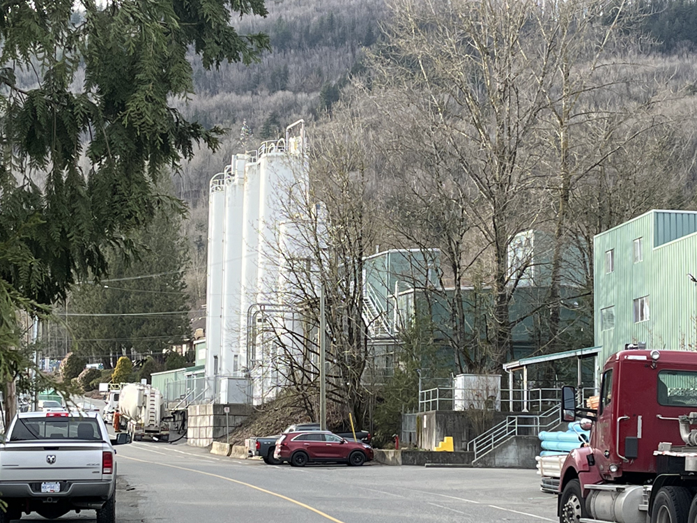 A street has residences on one side, and a PVC piping factory on the opposite side. There is an industrial building, with silos. There’s a treed hill in the background.