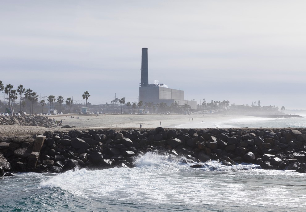 A factory with a smokestack is located near a beach, which has big waves crashing.