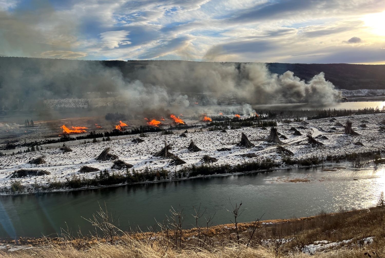 Amidst a snowy rural landscape, many burning piles of wood send up smoke.