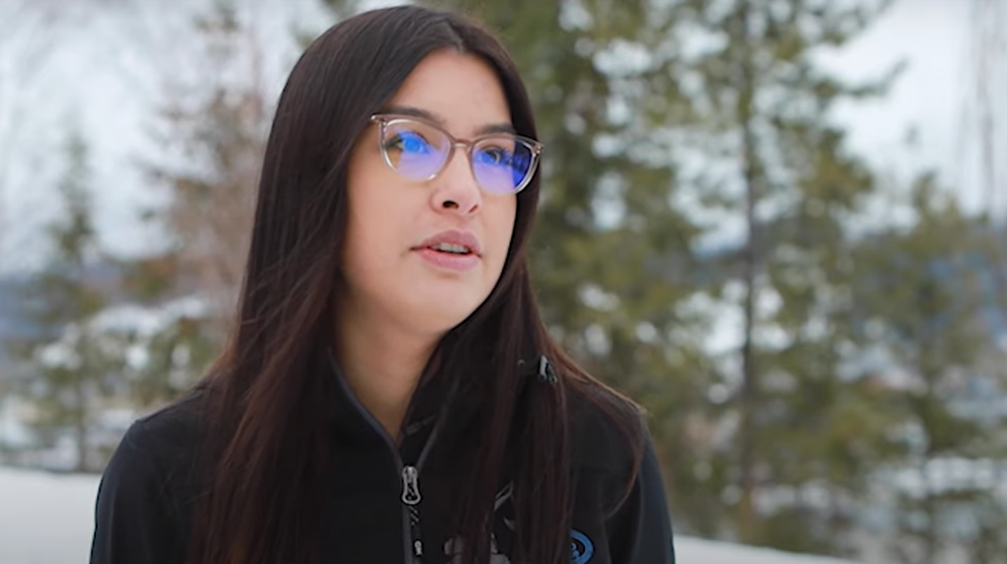 A young woman with black hair and glasses speaks with trees and snow out of focus behind her.