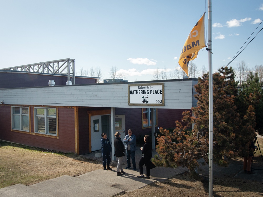 An “Every Child Matters” flag flies on a flag pole outside a one-storey grey and brown building with wooden siding. Four people gather in front of the building.