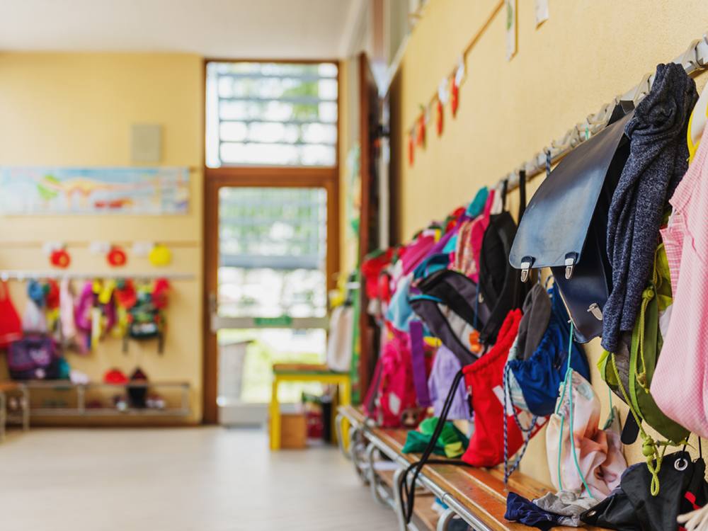 Coat hooks and a bench in a corner of a classroom that opens onto an outdoor setting. There are children’s clothes and bookbags hanging on the hooks.
