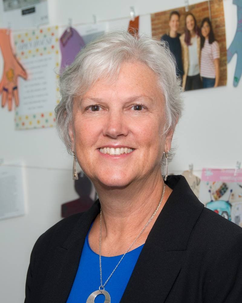 A woman with grey hair and earrings, wearing a bright blue shirt and a blazer, smiles at the camera.