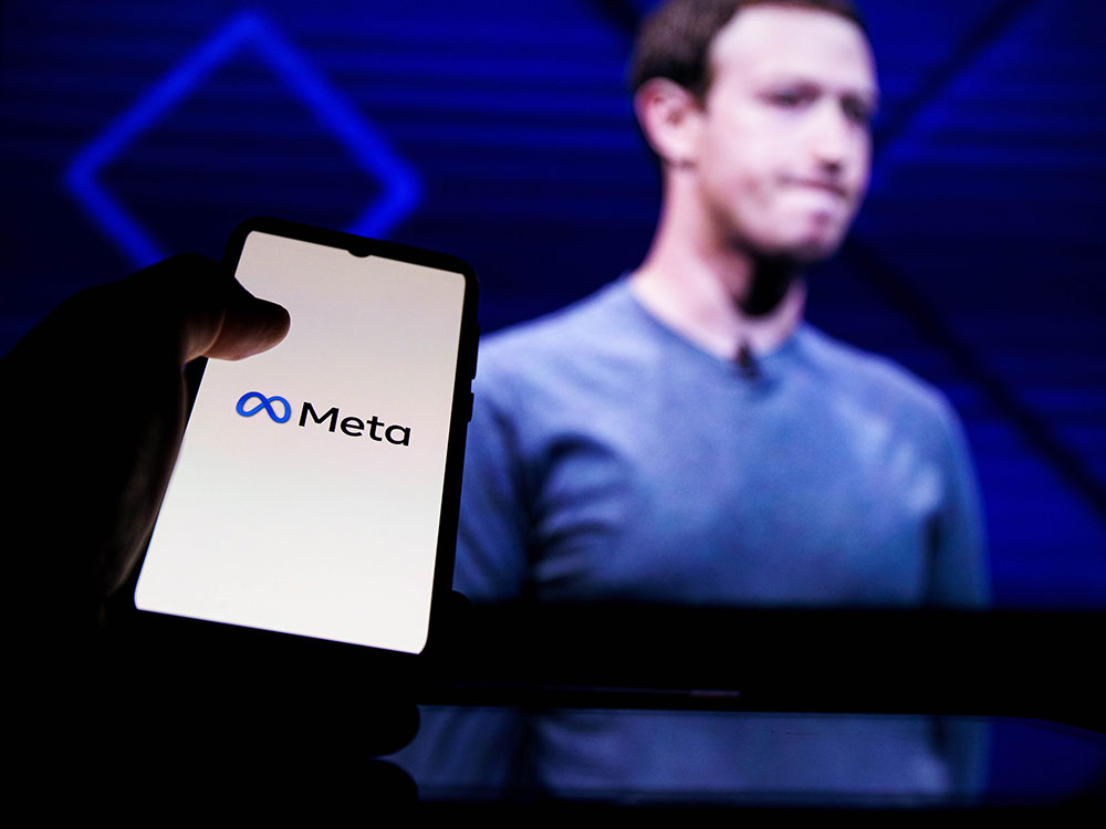 The image shows a hand holding a cellphone with Meta on the screen, with a blurry image of Mark Zuckerberg in the background.