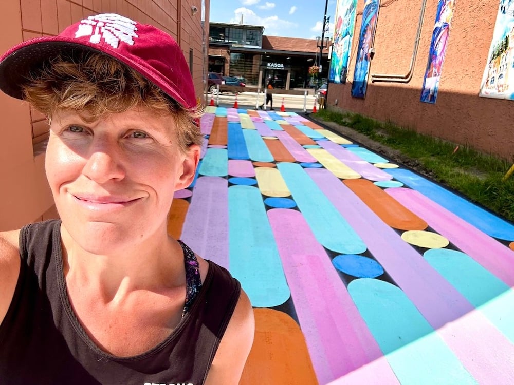 Janis Irwin stands to the left of the frame in a black tank top and red ball cap, smiling with her mouth closed. To her right and behind her is a colourful mural in which the laneway is painted with oblong pastel shapes in pink, blue, yellow and orange. It’s a summer day and a terra cotta wall with artwork on it lines the lane.