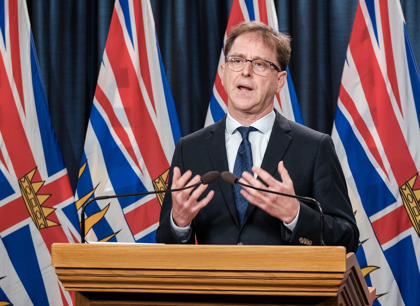 A man in a suit speaks at a conference in front of multiple flags for the Canadian province of British Columbia.