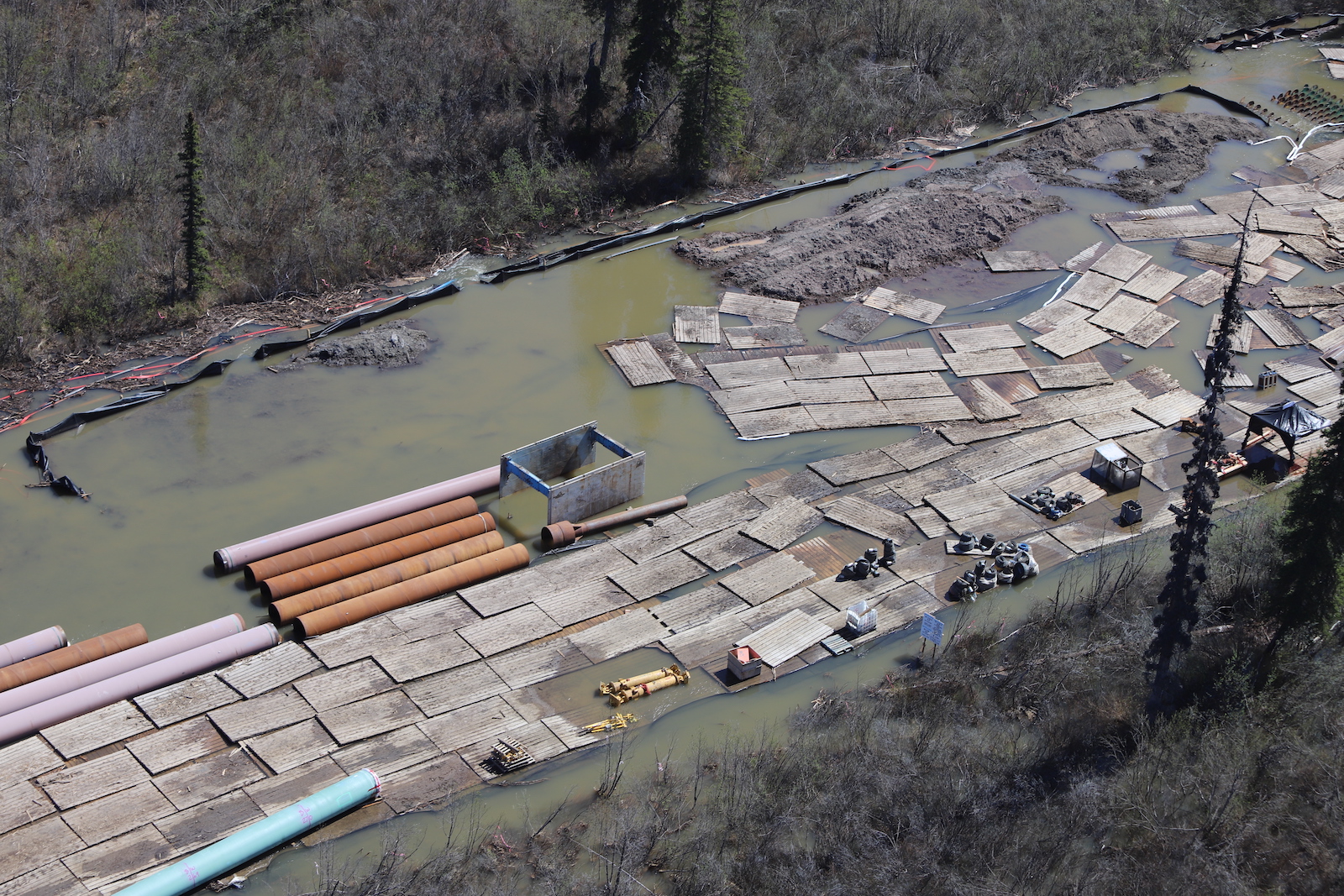 An aerial photograph shows dozens of sheets of plywood, heavy machinery and sections of metal pipe surrounded by brown water.