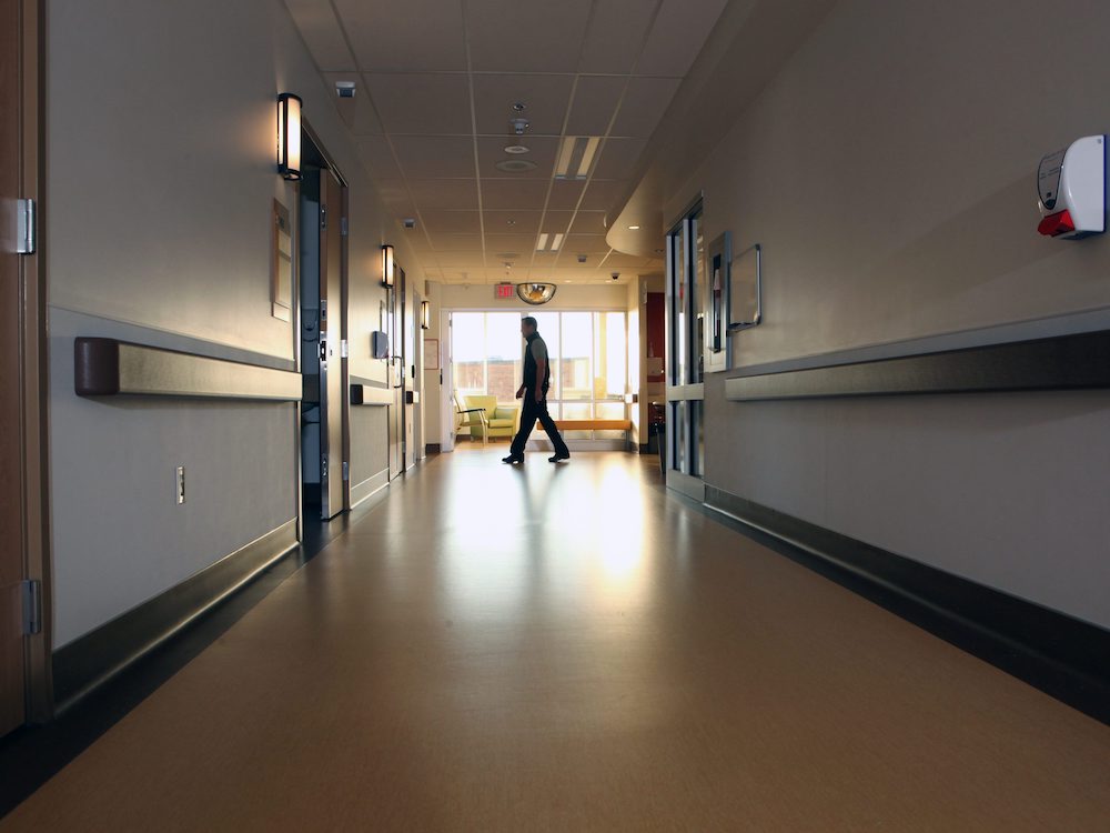 A dimly lit hospital corridor has light brown floors and grey and beige walls. At the end of the hallway is a bright set of windows. A man in a fleece vest is walking by.