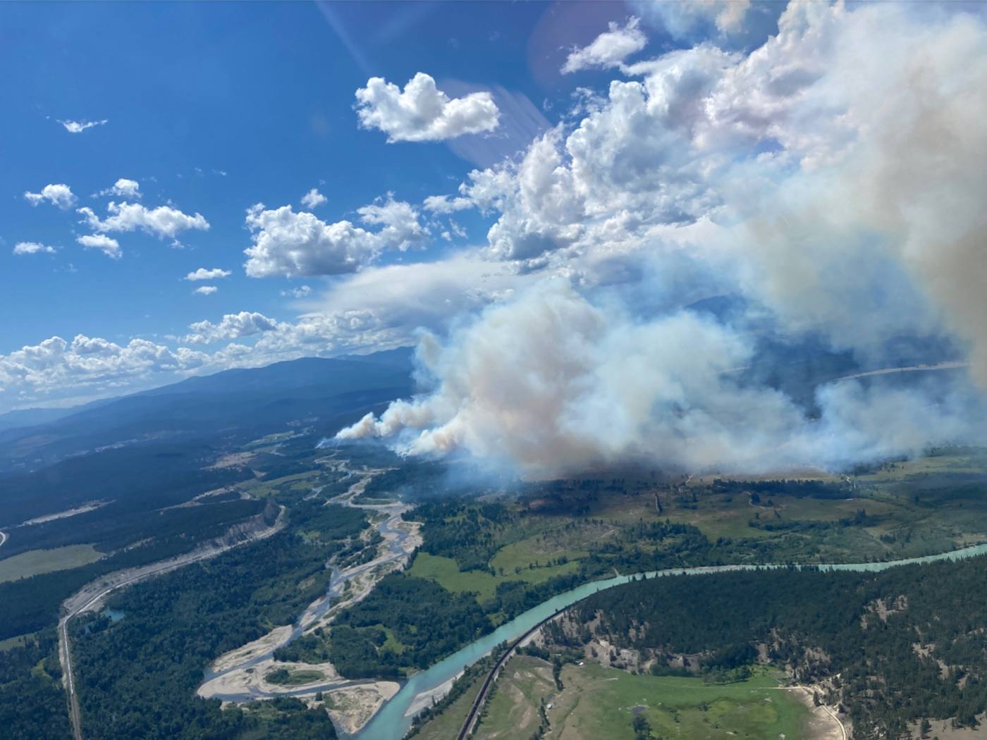 A large plume of smoke trails back through the sky towards the camera, over a treed landscape with a river.