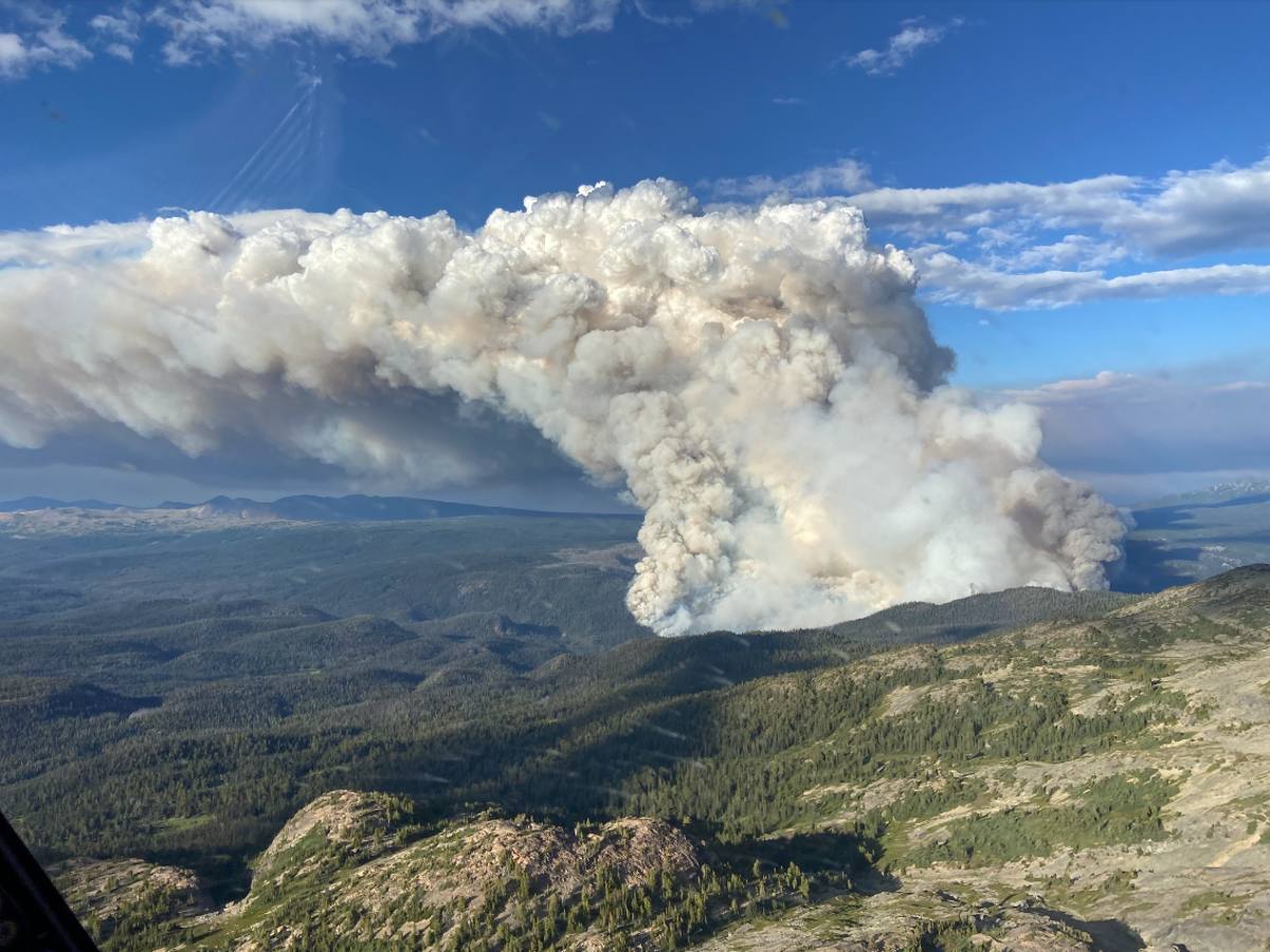 A massive plume of smoke rolls out away from the camera, over hilly treed terrain.