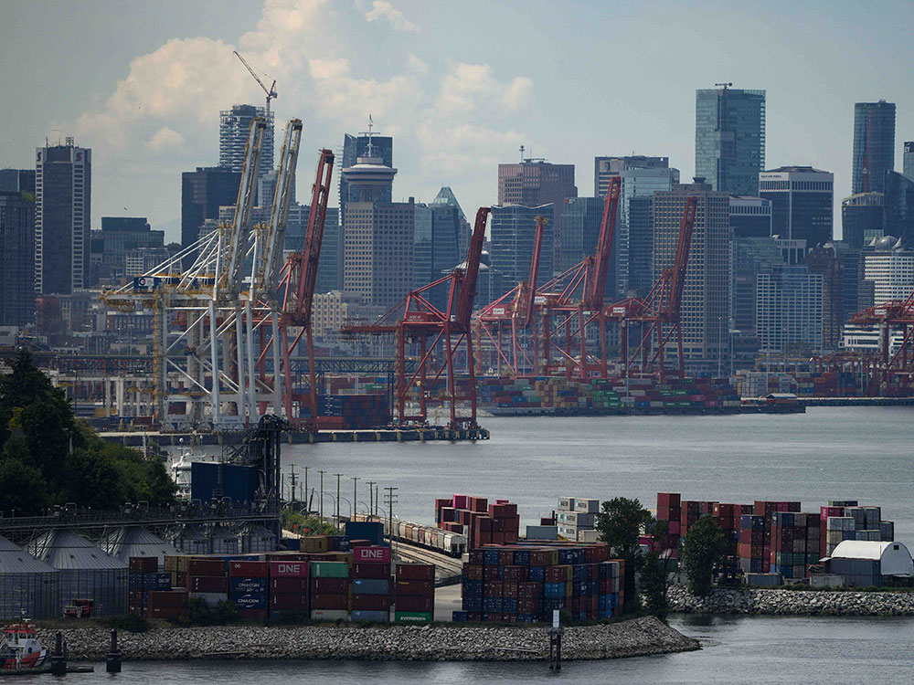 Shipping containers are stacked in the foreground and a row of tall gantry cranes loom over the docks, with Vancouver's skyline in the background.