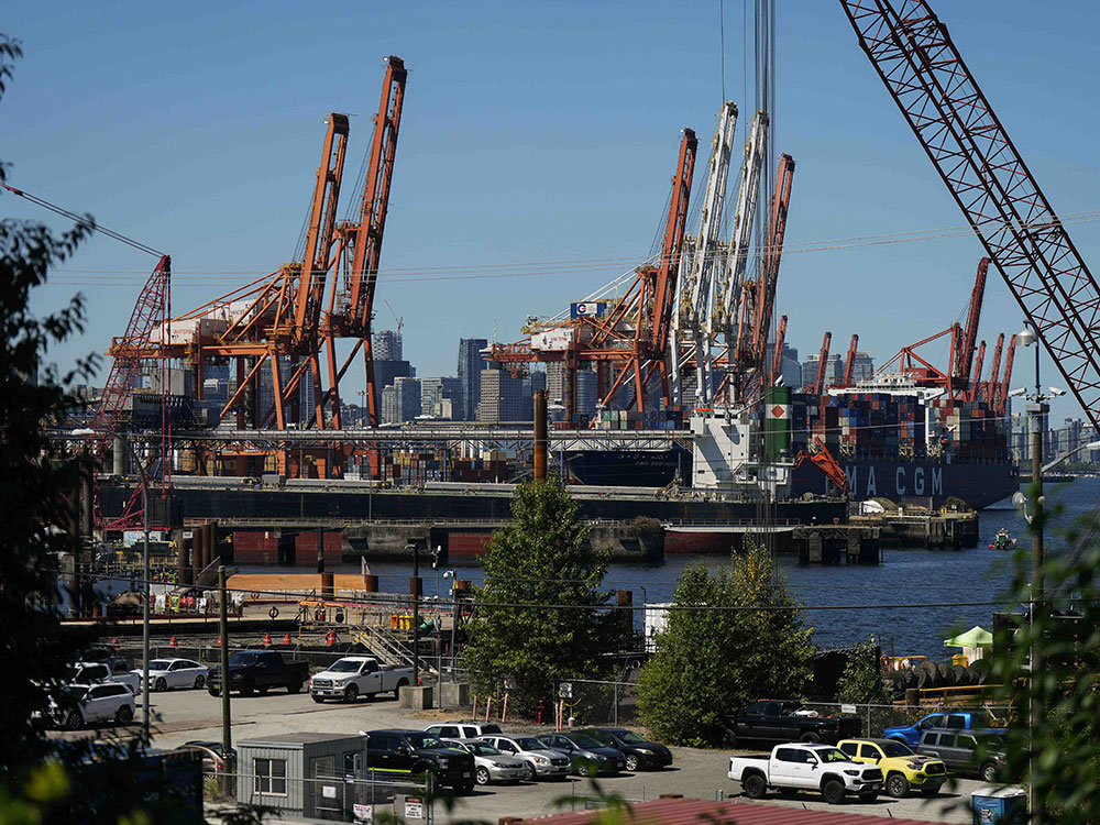 Gantry cranes loom over a container ship, with the Vancouver skyline in the background.