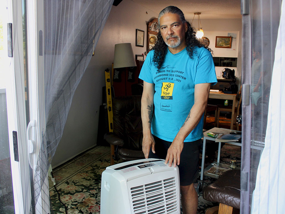 Ryan Le Neal, wearing a blue T-shirt and black shorts and with long hair and a beard, shows the portable air conditioner he uses to cool his apartment.
