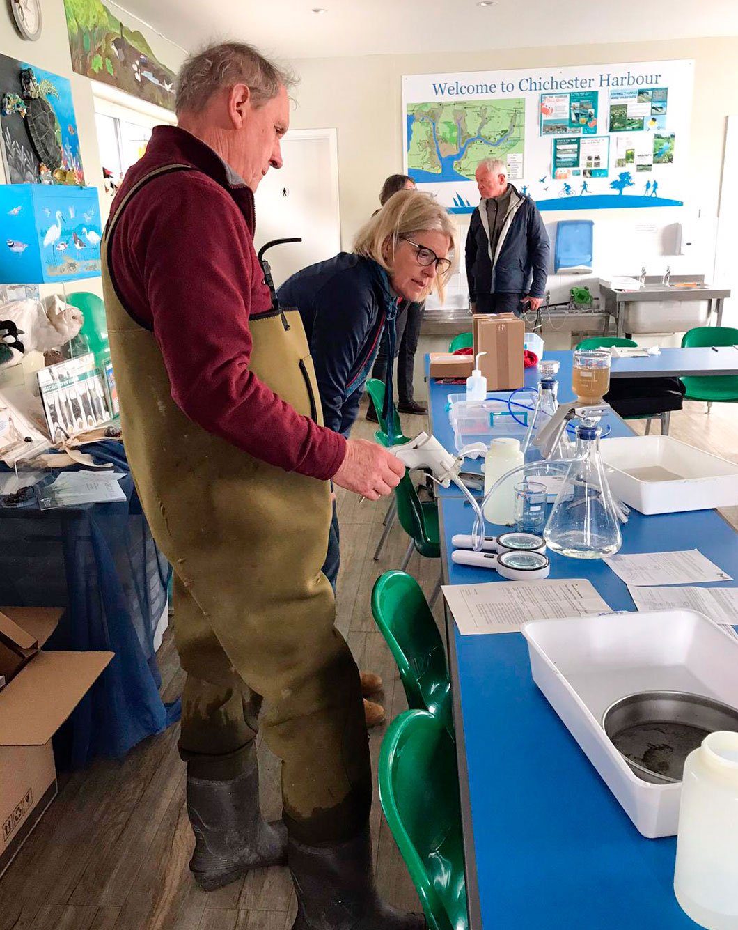 Two people stand at a table with beakers and other equipment. One person is wearing hip waders.