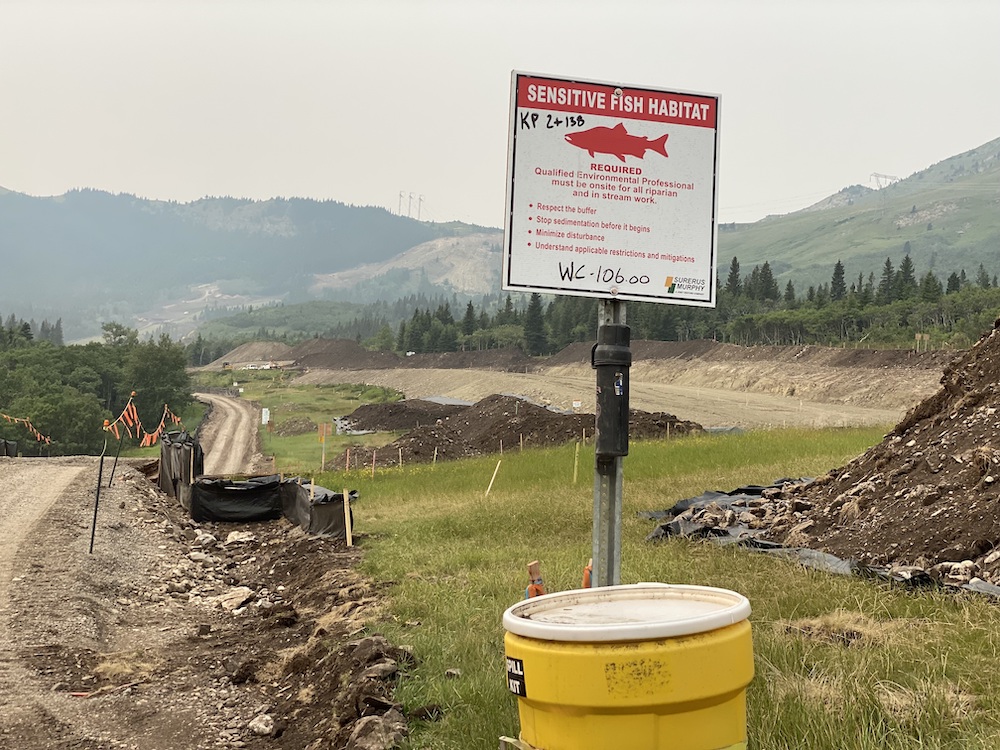 In the foreground, a red and white sign reads “sensitive fish habitat.” In the background, there are large piles of dirt and a newly built access road that is marked with orange flagging tape.