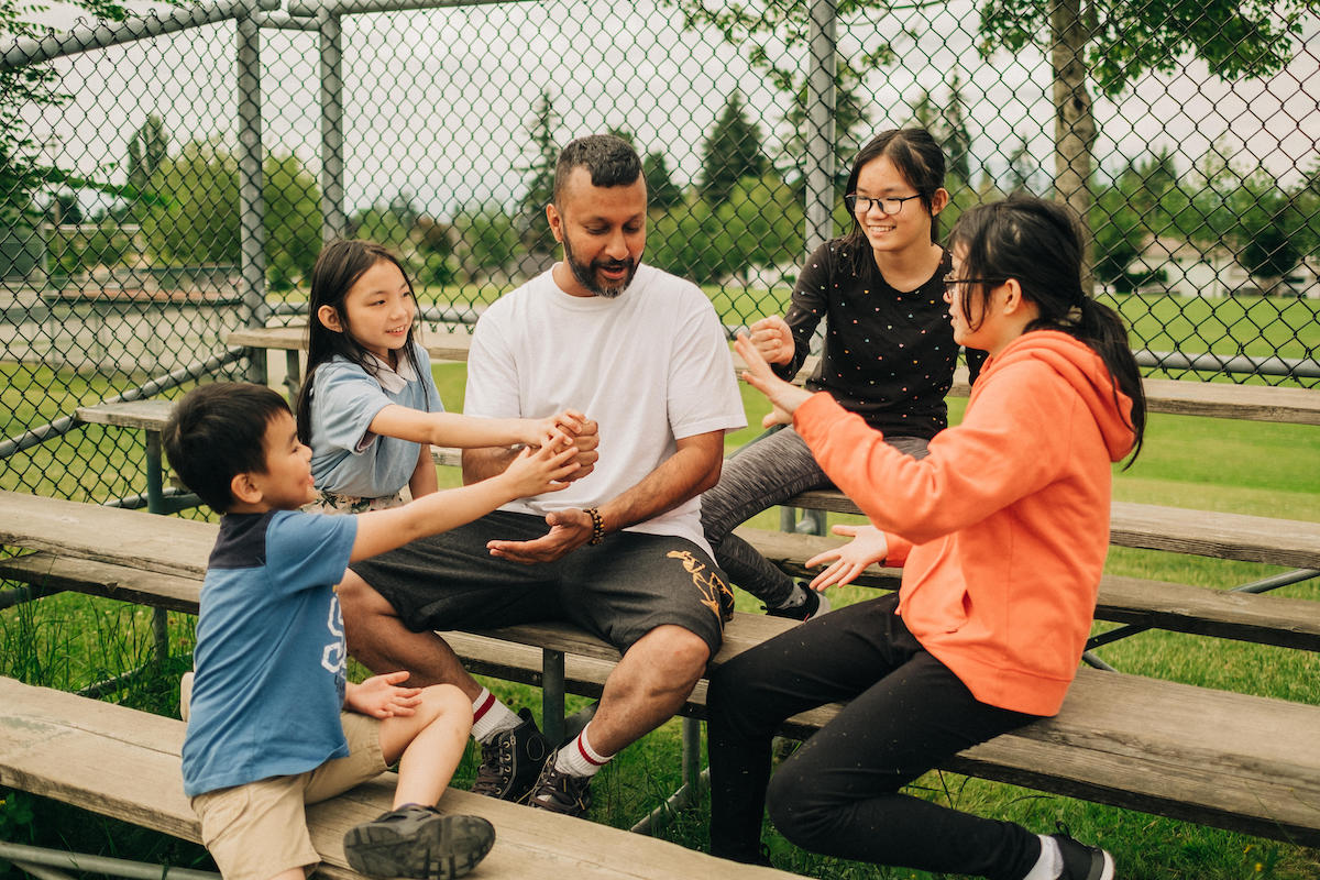 Four kids sit on bleachers outside with a camp counsellor, playing a game.