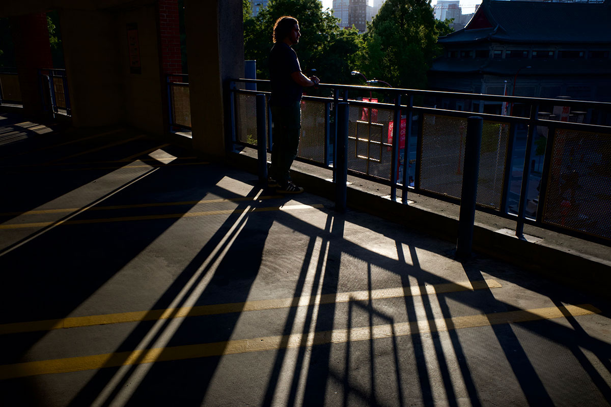 A man stands on a balcony, looking out over a street. Long shadows are cast behind him.