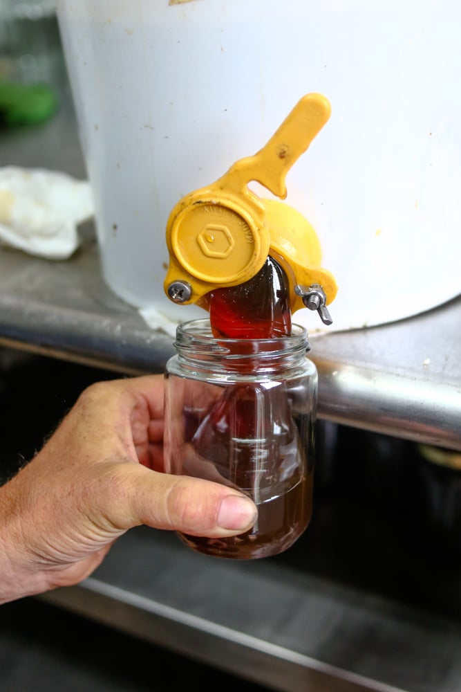 A white bucket with a tap attached to it, from which a man is pouring honey into a jar.
