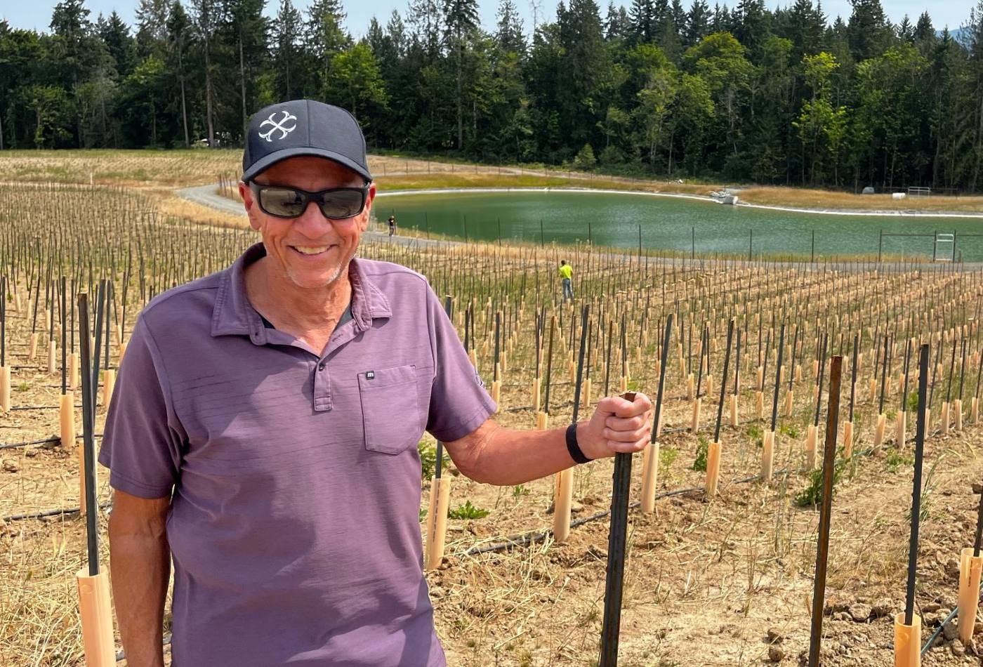 A late middle-aged white man in ball cap and sunglasses and a short sleeved collared shirt stands amidst young grape vines growing in rows, with a pond and strip of forest behind him. 