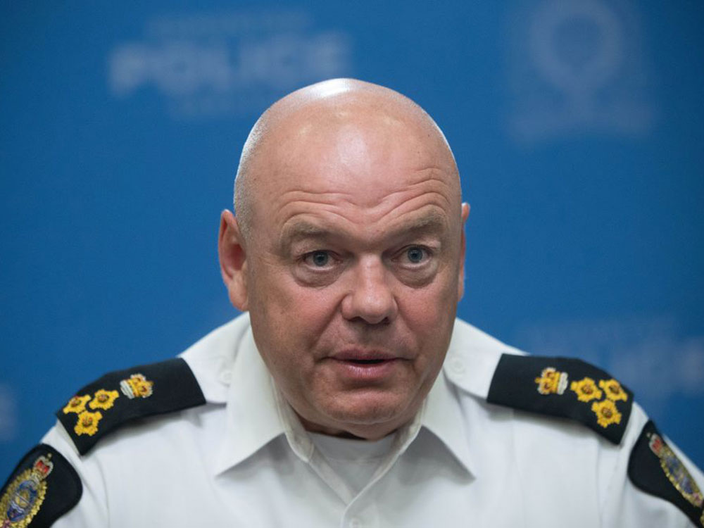 A photograph of a bald man in a white collared shirt with police badges sewn over the shoulders. He is looking directly at the camera in front of a blurred blue background.