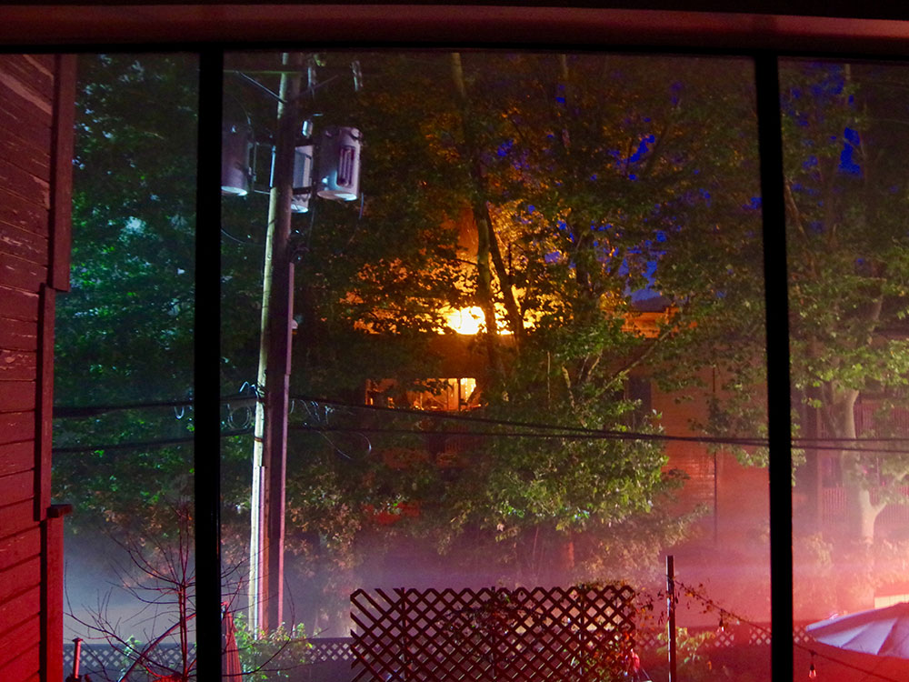 Through leafy green tree branches, the intense yellow glow of a fire in a wooden apartment building can be seen.