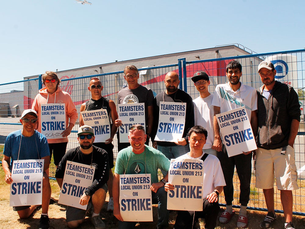 Eleven striking workers pose for a photo in front of the Coca Cola factory. Many hold signs saying ‘Teamsters Local Union 213 on Strike.’
