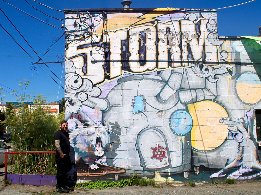 A man dressed in black with a backwards baseball cap stands next to a mural that reads ‘STORM’ on an old brick building. There are power lines and reaching from the building across a blue sky, and overgrown weeds in the background.