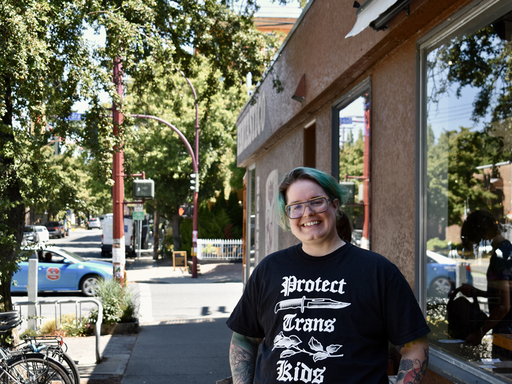 Sloane Chomeakwich stands outside Caffe Fantastico on a sunny day wearing a shirt that says ‘Protect Trans Kids.’