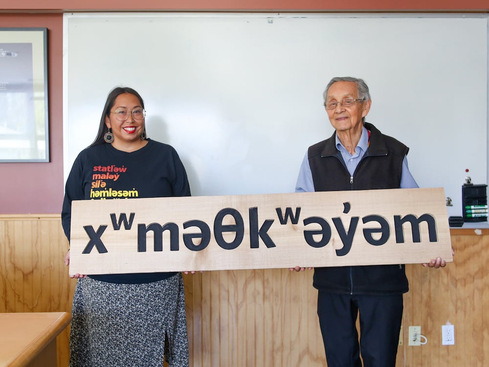 An Indigenous woman in her 30s and an Indigenous man in his 80s hold up a wooden sign that displays Musqueam’s name in the North American Phonetic Alphabet.