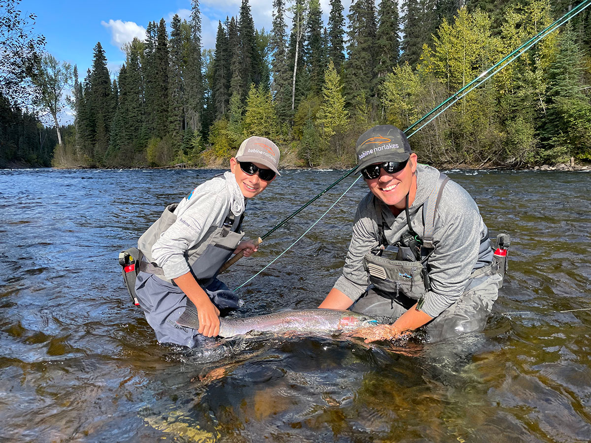 A youth and a man stand in a river holding a large fish and smiling at the camera. The youth also holds a fishing rod. 