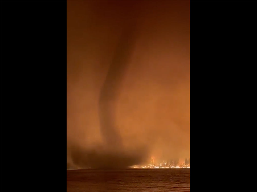 A tornado rises from a lake ground into the sky, with fires in the background.