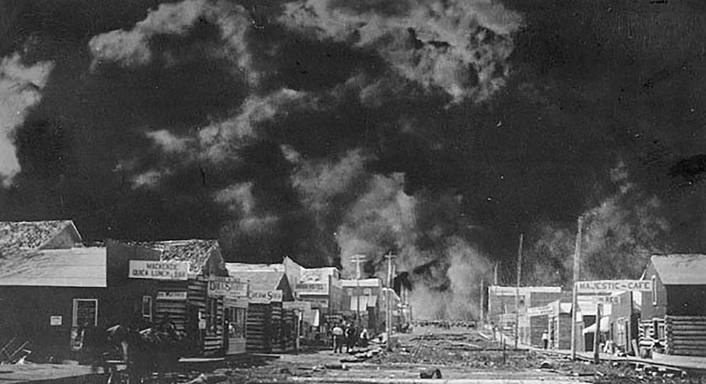 A dirt street with wood buildings on both sides and a massive wall of smoke behind.