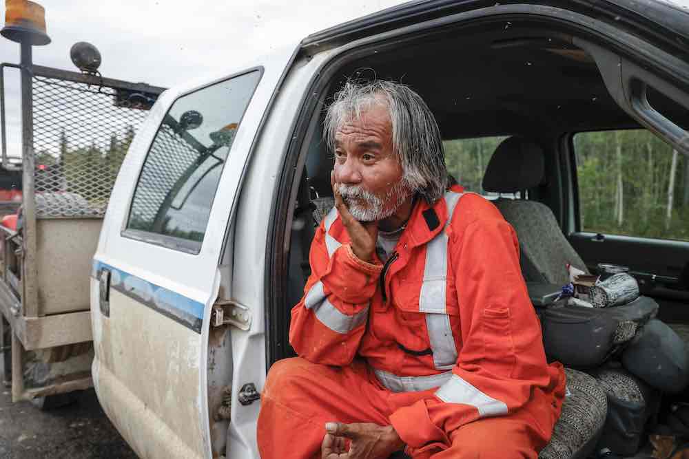 A man with shaggy greying hair and a beard sits with his hand cupping his face. He is wearing an orange construction jumpsuit and sitting on the grey upholstered seat of a dirty white cargo van with his body facing out the door.