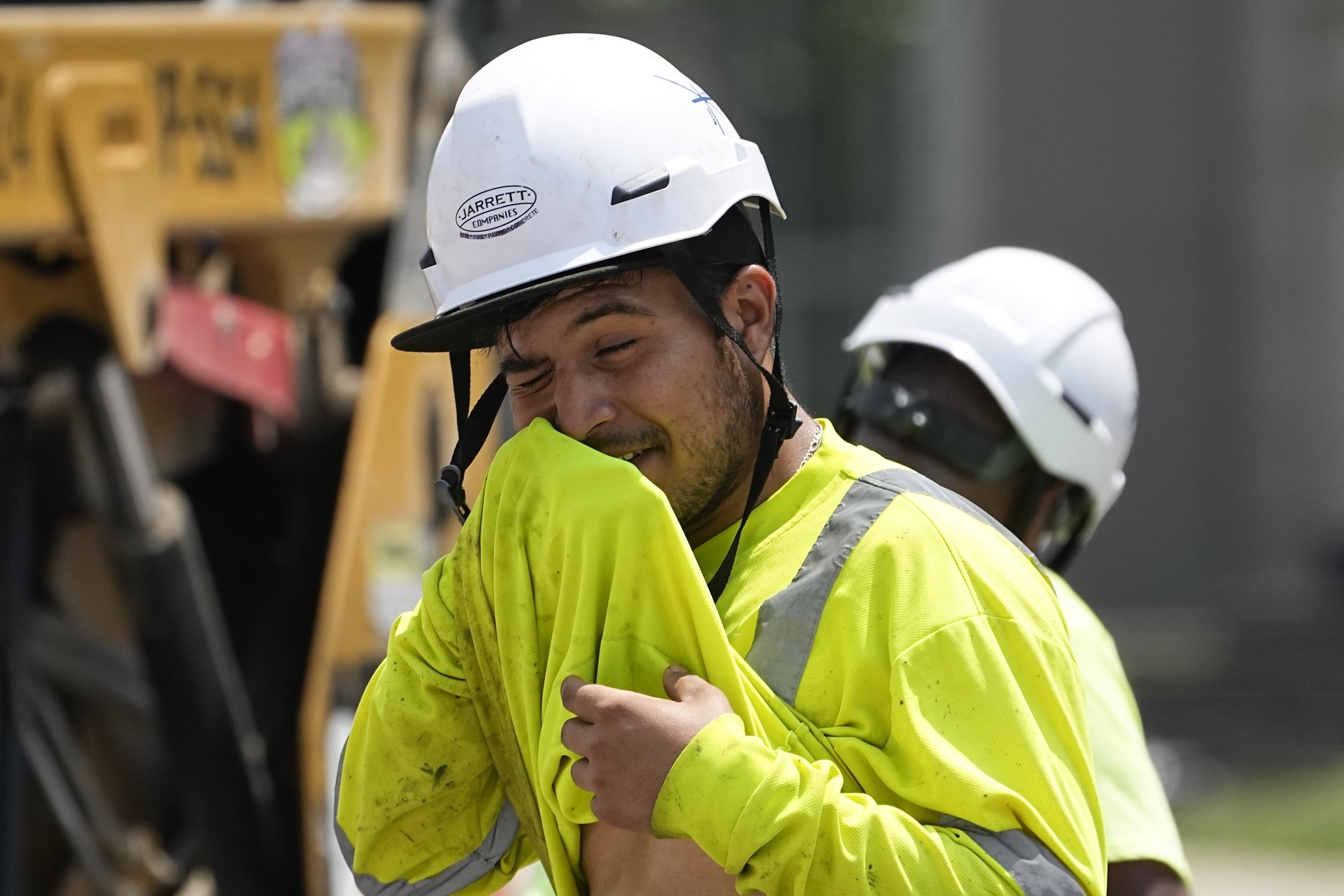 A worker in safety gear and a white helmet wipes his face in the heat.
