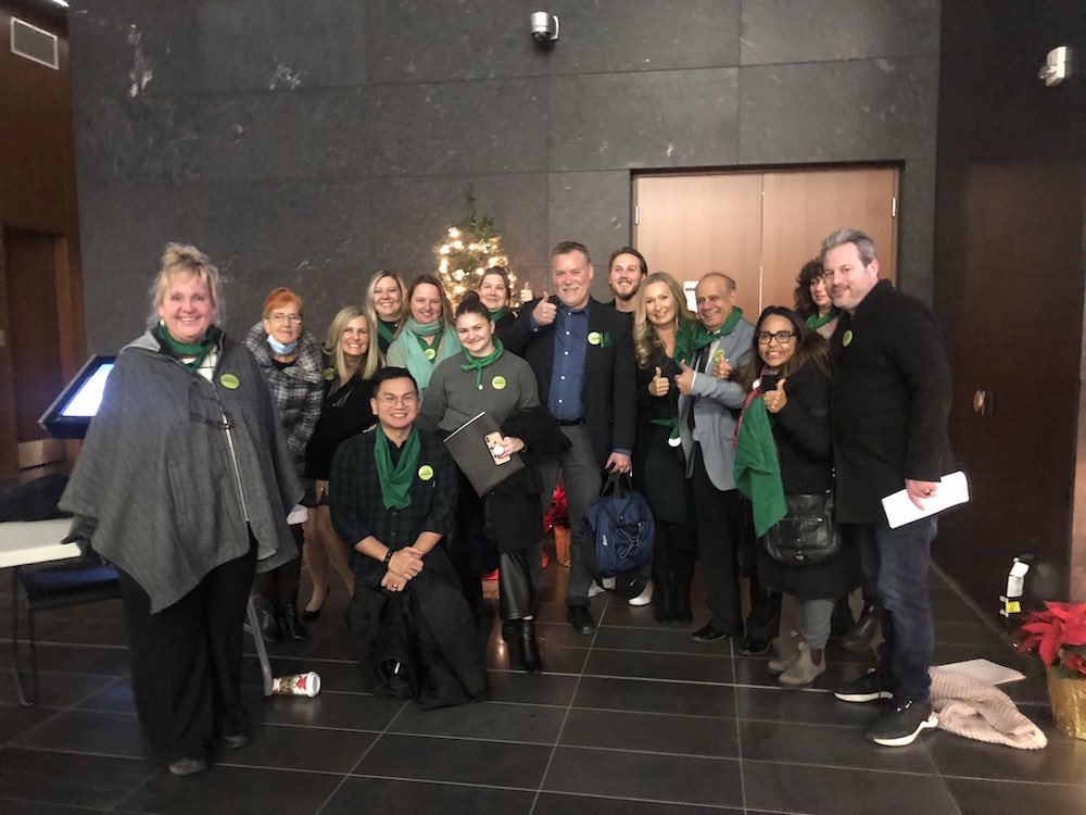 A crowd of people looking at the camera in the lobby of a nice administrative building with a Christmas tree in the back. A man in the centre is holding a thumb up.