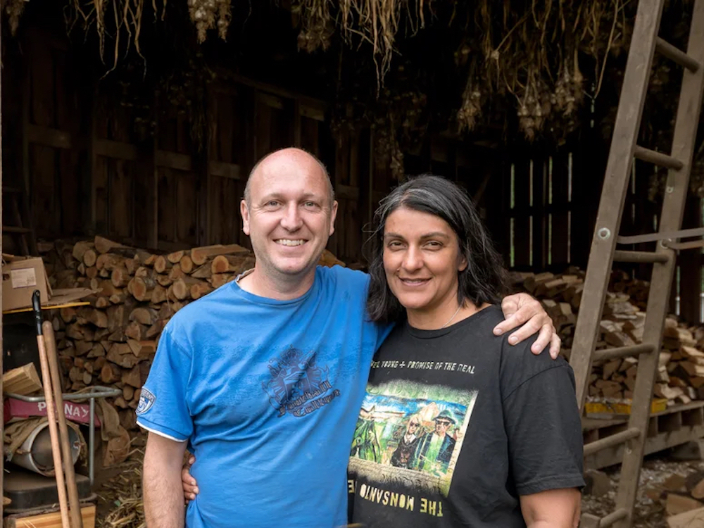 Two people standing in front of a woodshed smile at the camera.