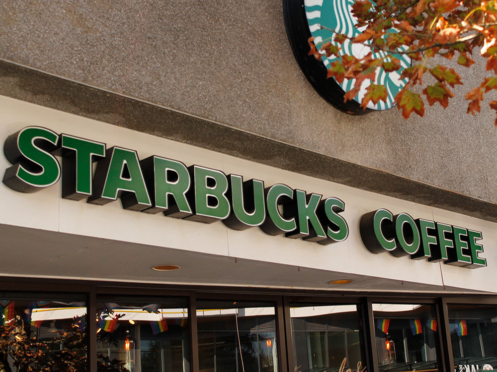 The front of a Starbucks outlet, with the words 'Starbucks Coffee' and the familiar logo. Fall leaves are in the foreground over the logo.