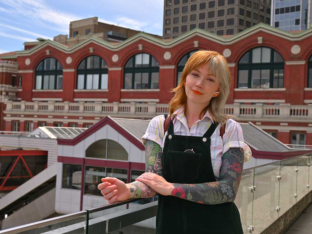 A woman wearing corduroy overalls with sleeve tattoos stands near a railing. Waterfront station is in the background.